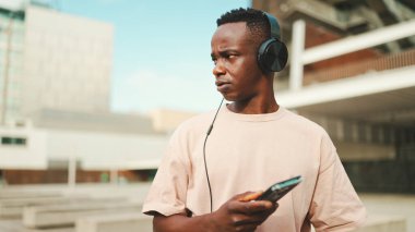 Üniversitenin dışında oturan genç Afrikalı öğrenci kulaklık takıyor, telefon kullanıyor, müzik dinliyor.