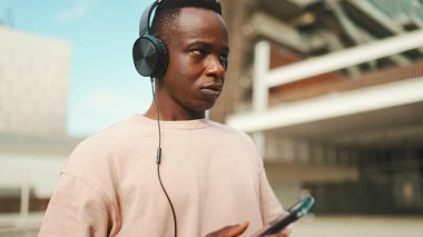 Üniversitenin dışında oturan genç Afrikalı öğrenci kulaklık takıyor, telefon kullanıyor, müzik dinliyor.