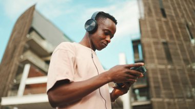 Üniversitenin dışında oturan genç Afrikalı öğrenci kulaklık takıyor, telefon kullanıyor, müzik dinliyor.