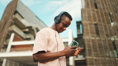 Üniversitenin dışında oturan genç Afrikalı öğrenci kulaklık takıyor, telefon kullanıyor, müzik dinliyor.