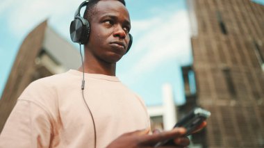 Üniversitenin dışında oturan genç Afrikalı öğrenci kulaklık takıyor, telefon kullanıyor, müzik dinliyor.