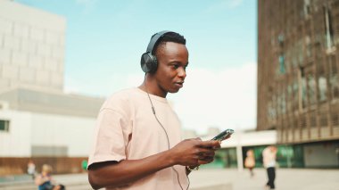 Üniversitenin dışında oturan genç Afrikalı öğrenci kulaklık takıyor, telefon kullanıyor, müzik dinliyor.