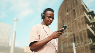 Üniversitenin dışında oturan genç Afrikalı öğrenci kulaklık takıyor, telefon kullanıyor, müzik dinliyor.