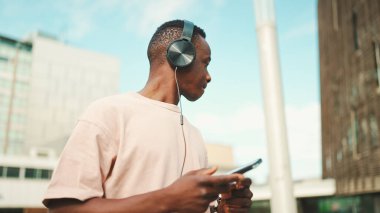 Üniversitenin dışında oturan genç Afrikalı öğrenci kulaklık takıyor, telefon kullanıyor, müzik dinliyor.