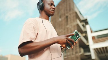 Üniversitenin dışında oturan genç Afrikalı öğrenci kulaklık takıyor, telefon kullanıyor, müzik dinliyor.