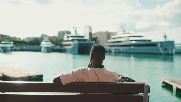 Young African Guy Relaxes Sitting Bench Port Yachts Ships Background — Φωτογραφία Αρχείου