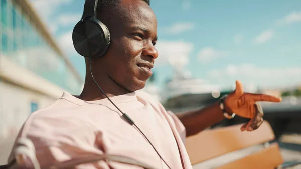 Smiling young african guy relaxes sitting on bench in the port, listens to music in headphones, sings along