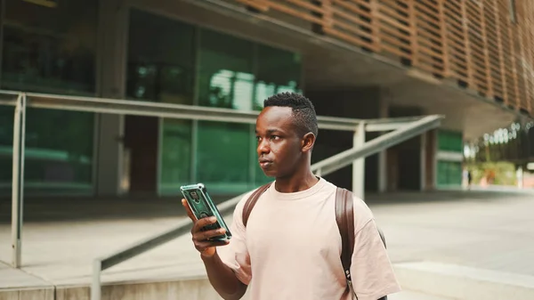Young african student walks down the stairs outside of the university, using cellphone