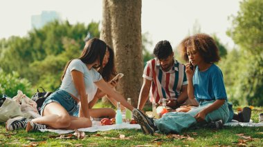 Kıvırcık saçlı, çizgili tişörtlü, parkta oturmuş, yaz günü dışarıda piknik yapan arkadaşlarıyla konuşan genç bir adam.