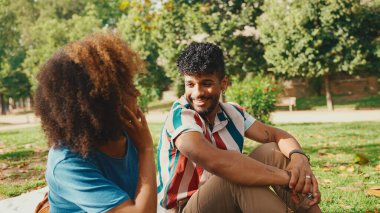 Kıvırcık saçlı, çizgili tişörtlü, parkta oturmuş, yaz günü dışarıda piknik yapan arkadaşlarıyla konuşan genç bir adam.