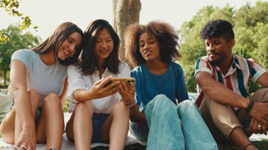 Mutlu, çoklu etnik çeşitlilikte gülümseyen gençler yaz günü açık havada piknik yapıyorlar. Bir grup arkadaş piknikte dinlenirken konuşuyor, cep telefonu kullanıyor.