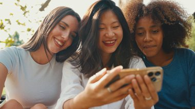 Happy, smiling multiethnic young people at picnic on summer day outdoors. Group of friends use cellphone, watch videos, social networks