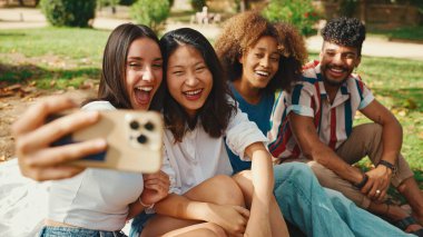Happy multiethnic young people are looking at taking selfie on summer day outdoors. Group of smiling friends posing at smartphone camera