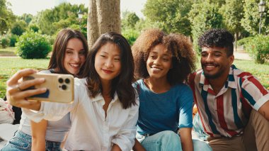 Happy multiethnic young people are looking at taking selfie on summer day outdoors. Group of smiling friends posing at smartphone camera