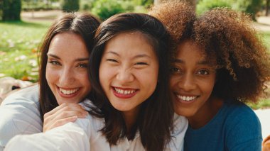 Close-up of happy multiethnic young women posing on cellphone camera outdoors. Girls look at the camera and laugh merrily and take selfie