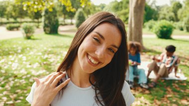 Parktaki kameraya poz veren uzun kahverengi saçlı, gülümseyen genç bir kadının yakın çekimi. Yaz günü açık havada piknik. Arkadaşları arka planda bulanık oturuyor.