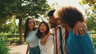 Mutlu çoklu etnik çeşitlilikte genç insanlar yaz günü açık havada kucaklaşarak yürürler. Bir grup arkadaş, şehir parkında yürürken neşeyle konuşuyor ve gülüyorlar.