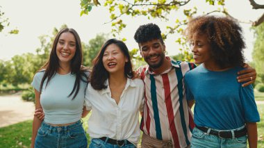Happy multiethnic young people walk embracing on summer day outdoors. Group of friends are talking and laughing merrily while walking along path in city park
