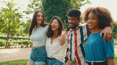 Happy multiethnic young people walk embracing on summer day outdoors. Group of friends are talking and laughing merrily while walking along path in city park