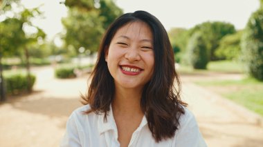 Close-up young Asian smiling woman with long brown hair wearing white t-shirt posing for the camera in the park . The girl opens her eyes and smiles at the camera