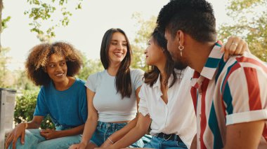 Happy multiethnic young people talking while sitting on park bench on summer day outdoors. Group of friends talking and laughing merrily in city park