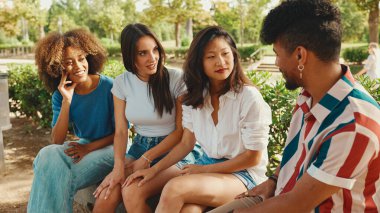 Yaz günü açık havada parkta bankta oturan mutlu çoklu etnik gruptan gençler konuşuyor. Şehir parkında bir grup arkadaş neşeyle konuşuyor ve gülüyor.