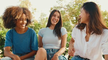 Happy multiethnic young women talking while sitting on park bench on summer day outdoors. Group of girls talking and laughing merrily in city park