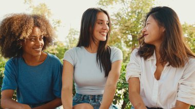 Happy multiethnic young women talking while sitting on park bench on summer day outdoors. Group of girls talking and laughing merrily in city park