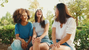Yaz günü açık havada parkta bankta oturan mutlu çoklu etnik kökenli genç kadınlar konuşuyor. Şehir parkında bir grup kız neşeyle konuşuyor ve gülüyor.