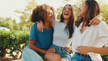 Happy multiethnic young women talking while sitting on park bench on summer day outdoors. Group of girls talking and laughing merrily in city park