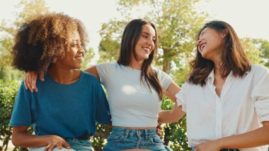 Happy multiethnic young women talking while sitting on park bench on summer day outdoors. Group of girls talking and laughing merrily in city park