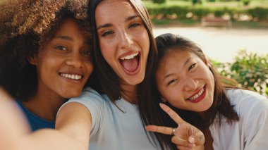 Happy, lovely multiethnic young women posing on cellphone camera on summer day outdoors. group of friends hugging each other smiling at the camera while standing on path in the park