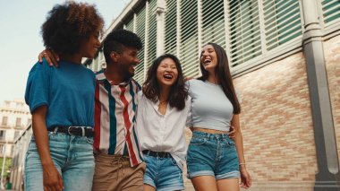 Happy multiethnic young people walk embracing on summer day outdoors. Group of friends are talking and laughing merrily while walking along on the street