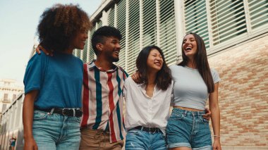 Mutlu çoklu etnik çeşitlilikte genç insanlar yaz günü açık havada kucaklaşarak yürürler. Bir grup arkadaş sokakta yürürken neşeyle konuşuyor ve gülüyorlar.