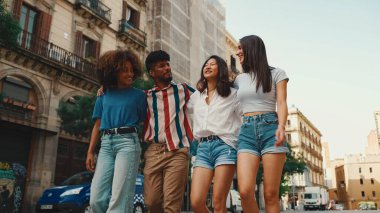 Mutlu çoklu etnik çeşitlilikte genç insanlar yaz günü açık havada kucaklaşarak yürürler. Bir grup arkadaş sokakta yürürken neşeyle konuşuyor ve gülüyorlar.