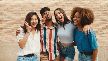 Happy, lovely multiethnic young people posing for the camera on summer day outdoors. Group of friends hugging tightly smiling at camera while standing on brick wall background