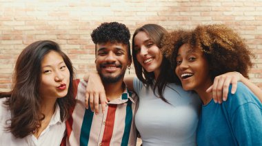 Happy, lovely multiethnic young people posing for the camera on summer day outdoors. Group of friends hugging tightly smiling at camera while standing on brick wall background