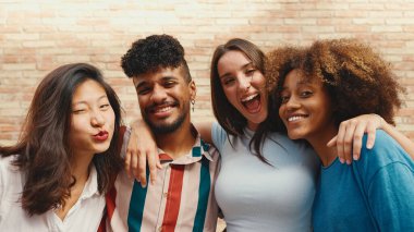 Happy, lovely multiethnic young people posing for the camera on summer day outdoors. Group of friends hugging tightly smiling at camera while standing on brick wall background