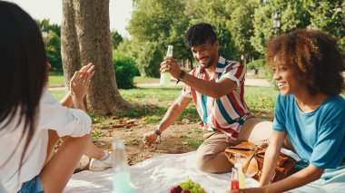 Yaz günü açık havada piknikte gülümseyen mutlu çokuluslu genç insanlar. Arkadaşlar hafta sonu eğleniyor, piknikte parkta dinleniyorlar.