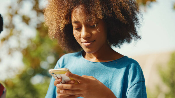 Close-up of young smiling woman with curly hair sitting in park having picnic with friends on summer day outdoors. Girl uses cellphone, browses social networks, photo, video