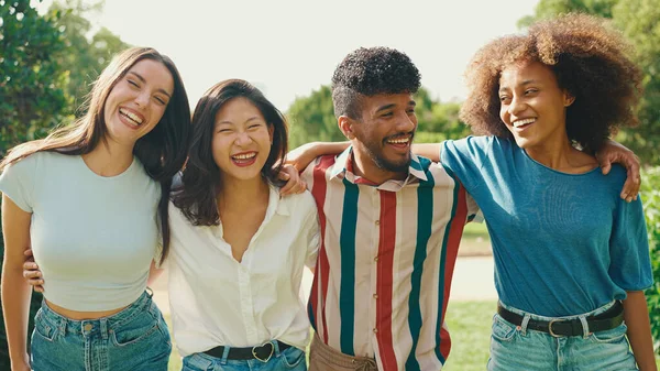 Happy multiethnic young people walk embracing on summer day outdoors. Group of friends are talking and laughing merrily while walking along path in city park