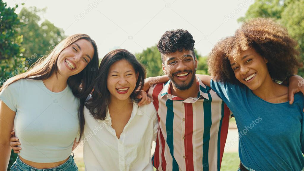 Happy multiethnic young people walk embracing on summer day outdoors. Group of friends are talking and laughing merrily while walking along path in city park