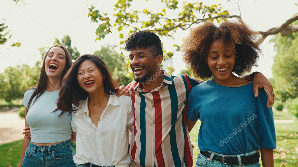 Happy multiethnic young people walk embracing on summer day outdoors. Group of friends are talking and laughing merrily while walking along path in city park