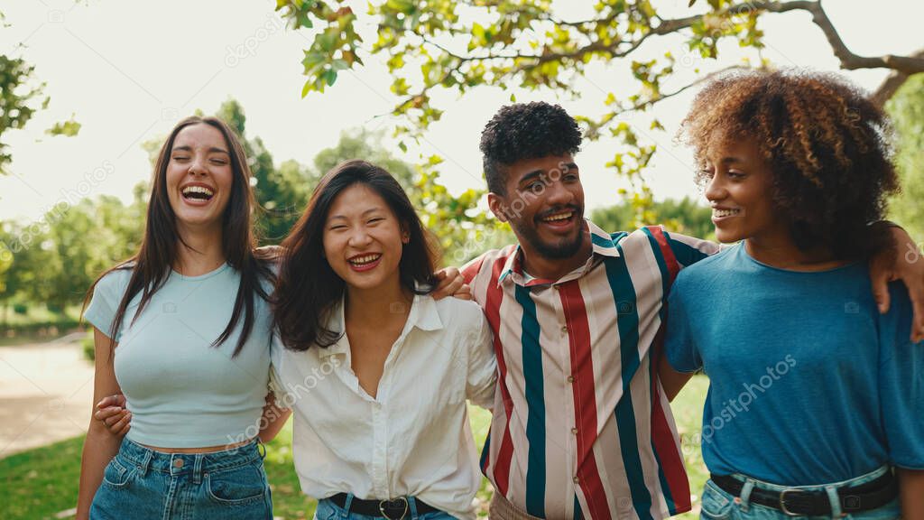 Happy multiethnic young people walk embracing on summer day outdoors. Group of friends are talking and laughing merrily while walking along path in city park