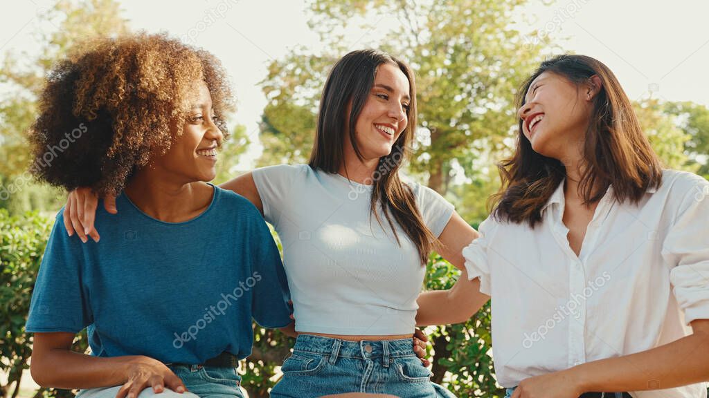 Happy multiethnic young women talking while sitting on park bench on summer day outdoors. Group of girls talking and laughing merrily in city park