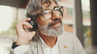 Clouse up portrait of mature businessman with beard in eyeglasses,wearing gray jacket makes conference video call on tablet pc. Middle aged man listening to audio materials in headphones.