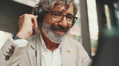 Gözlüklü ve kulaklıklı olgun bir iş adamı açık bir kafede oturup tablet kullanıyor.