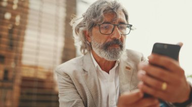 Gözlüklü, gri ceketli, sakallı, olgun bir işadamı cep telefonu kullanıyor. Orta yaşlı yönetici ofisin dışında otururken akıllı telefonuyla bilgi kaydırıyor.