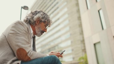 Gözlüklü, gri ceketli, sakallı, olgun bir işadamı cep telefonu kullanıyor. Orta yaşlı yönetici ofisin dışında otururken akıllı telefonuyla bilgi kaydırıyor.