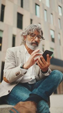 Gözlüklü, gri ceketli, sakallı, olgun bir işadamı cep telefonu kullanıyor. Orta yaşlı yönetici ofisin dışında otururken akıllı telefonuyla bilgi kaydırıyor.